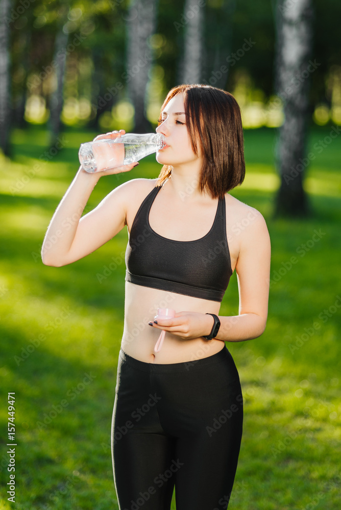 Attractive young sportsgirl finished her work out and now drinking water on the nature. She is standing on green grass in the forest Attractive young sportsgirl finished her work out and now drinking water on the nature. She is standing on green grass in the forest