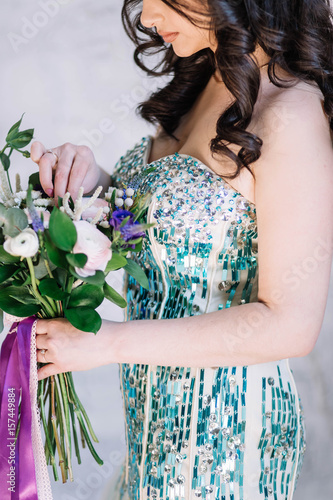 beautiful sexy Oriental girl with makeup black curls in a bright green sequined dress with purple and pink bouquet on his hand looking down profile on white gray background