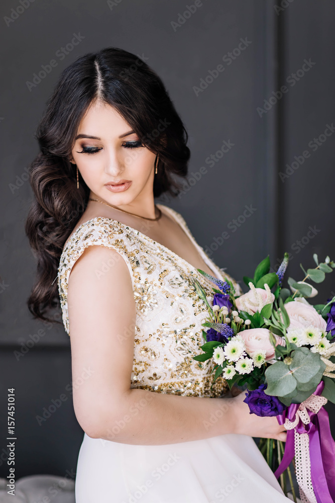 beautiful sexy Oriental girl brunette bride in white wedding dress with Golden sequins, a bouquet on gray background in Studio close-up, looks down, sitting on the edge of the sofa beautiful sexy Oriental girl brunette bride in white wedding dress with Golden sequins, a bouquet on gray background in Studio close-up, looks down, sitting on the edge of the sofa