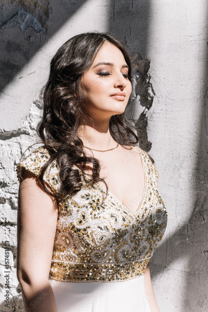 beautiful sexy Oriental girl in a white dress with a Golden glitter makeup long wavy hair standing on the hard sun with closed eyes smile close-up, portrait beautiful sexy Oriental girl in a white dress with a Golden glitter makeup long wavy hair standing on the hard sun with closed eyes smile close-up, portrait