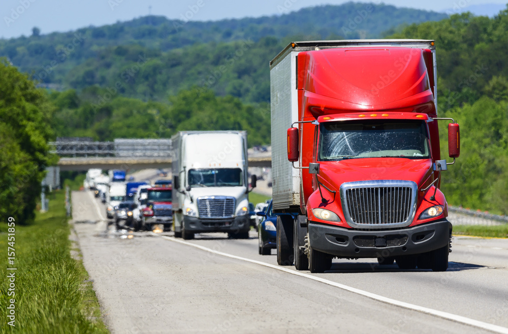 A red semi leads a line of traffic down an interstate highway in Tennessee.  Heat rising from the pavement gives background trucks and forest a cool shimmering effect.