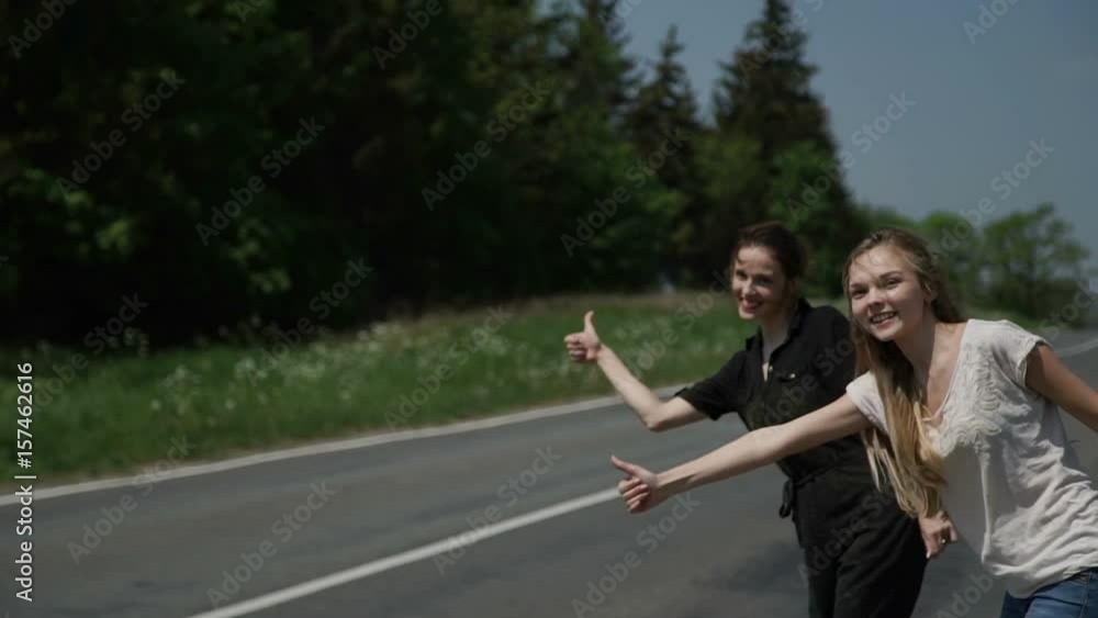 Two young women hitchhikers stand on the sidelines and ask to stop the passing cars Two young women hitchhikers stand on the sidelines and ask to stop the passing cars