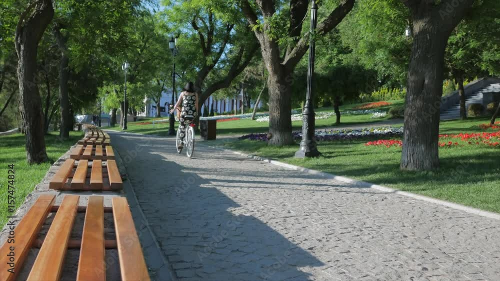 young beautiful woman wearing black overalls with sunflowers riding a vintage white bicycle in a park and passing by camera. Active people. Outdoors young beautiful woman wearing black overalls with sunflowers riding a vintage white bicycle in a park and passing by camera. Active people. Outdoors