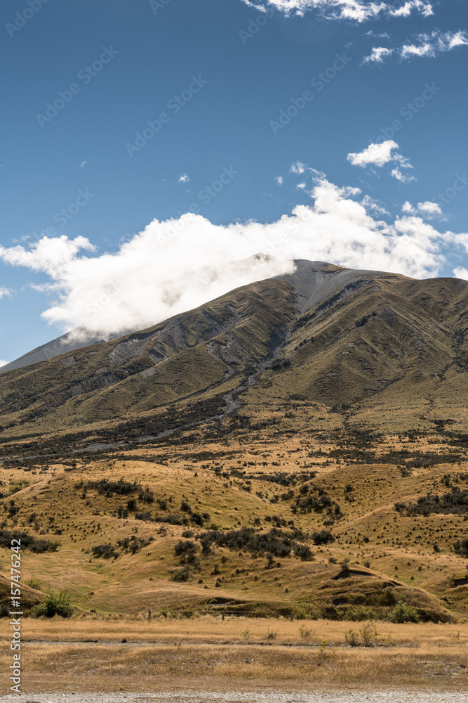 Middle Earth, New Zealand - March 14, 2017: Mountain at Middle Earth under blue cloudy sky. High desert scenery with sparse brown vegetation. Shale pebble flow off top. Middle Earth, New Zealand - March 14, 2017: Mountain at Middle Earth under blue cloudy sky. High desert scenery with sparse brown vegetation. Shale pebble flow off top.
