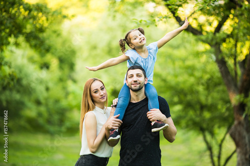 Father, mother and young daughter on piggyback with raised hands like flying on summer park background