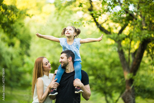 Father, mother and young daughter on piggyback with raised hands like flying on summer park background