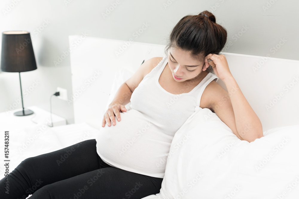 oung pregnant woman having a headache and she is lying on the bed holding her belly and forehead oung pregnant woman having a headache and she is lying on the bed holding her belly and forehead