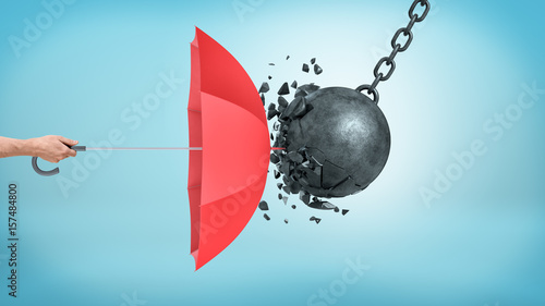 A male hand holding an open red umbrella which protects from a collision with a broken wrecking ball. photo