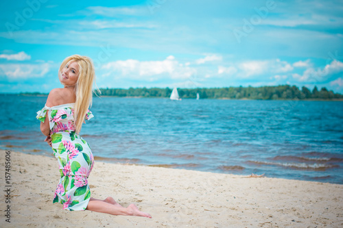 Beautiful blonde hair woman in romantic flowers dress near blue water and sand. Cute happy girl have fun on a nature