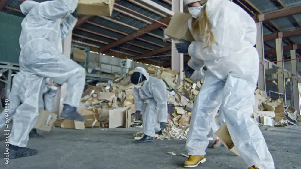 Low angle shot with PAN of excited garbage recycling factory workers in protective coveralls and masks throwing cardboard boxes at each other Low angle shot with PAN of excited garbage recycling factory workers in protective coveralls and masks throwing cardboard boxes at each other