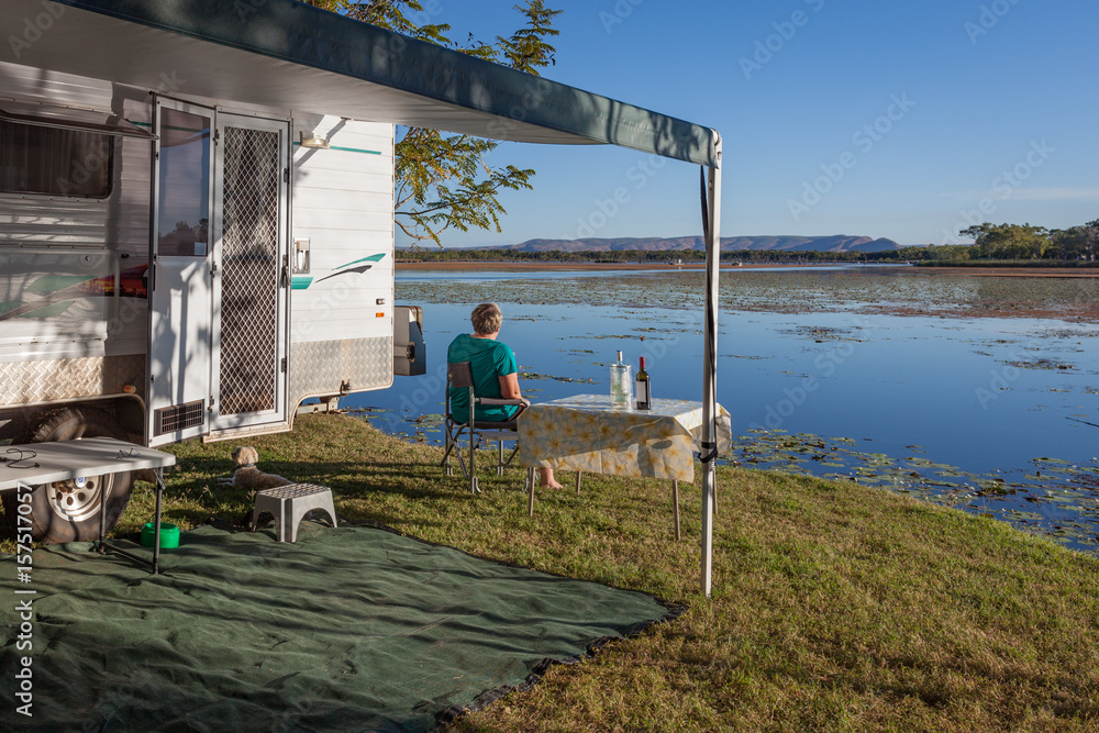 Retired mature lady sitting outside of a caravan with small dog enjoying a wine next to the Lilly filled Lake Kununurra.