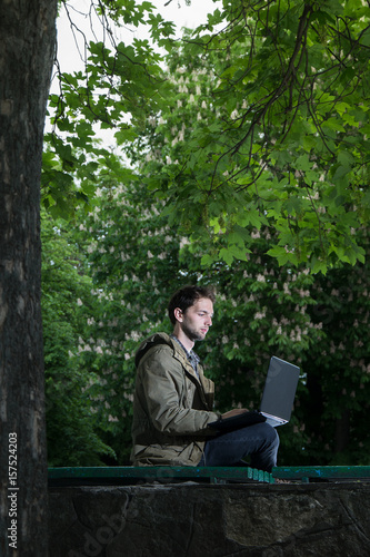 Young handsome student sitting on a wooden bench in the park with laptop computer.