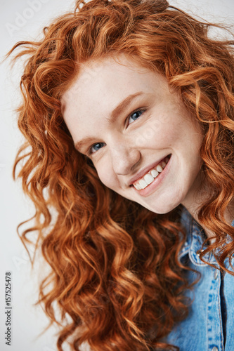 Close up of redhead beautiful girl with freckles smiling looking at camera over white background.