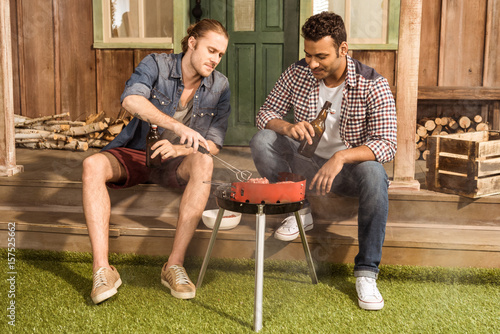 Young friends sitting on porch with beer bottles and and grilling meat