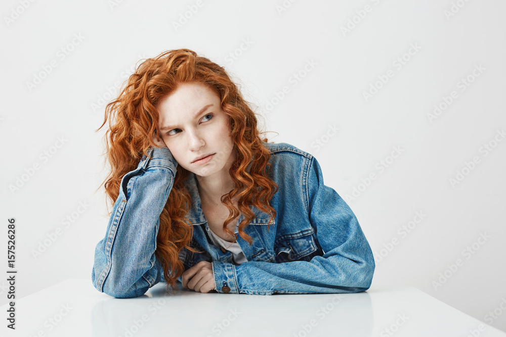 Displeased disappointed young girl with red curly hair sitting at table over white background Displeased disappointed young girl with red curly hair sitting at table over white background
