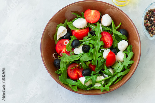 Close up of fresh salad with rucola, tomatoes olives, onion, mozzarella and spices in a bowl on a white stone background with copy space. Healthy food, Diet, Detox, Clean Eating or Vegetarian concept