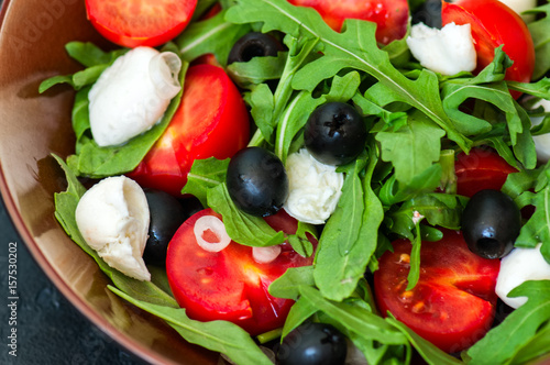 Close up of fresh salad with rucola, tomatoes, olives, onion, mozzarella and spices in a bowl on a black slate background with copy space. Healthy food, Diet, Detox, Clean Eating or Vegetarian concept