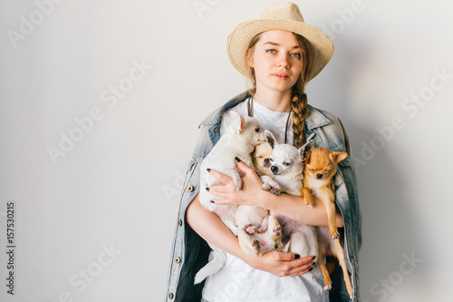 Young smiling happy hipster girl in straw hat, jeans jacket with russian style tress and upsweep holding in hands four chihuahua puppies dogs with funny emorional faces. Coiffure. Looking at camera. photo