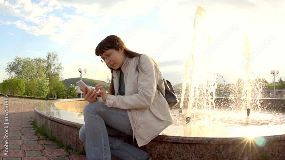 Young woman wearing glasses and a white jacket uses a phone while sitting on the edge of a city fountain during sunset. Young woman wearing glasses and a white jacket uses a phone while sitting on the edge of a city fountain during sunset.