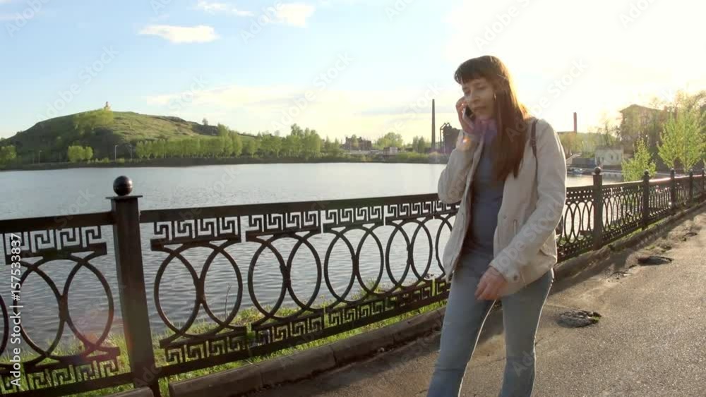 Young happy woman is walking along the river embankment in the city park talking on the phone on a bright sunny spring day. Young happy woman is walking along the river embankment in the city park talking on the phone on a bright sunny spring day.