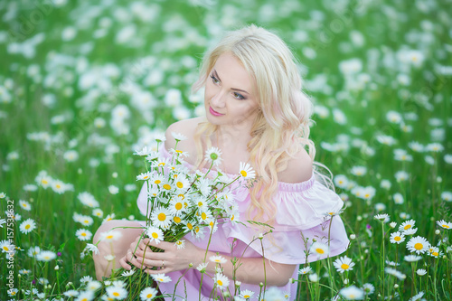 Attractive blonde in chamomile field. Young woman in wreath surrounded by chamomiles wearing stylish pink dress enjoying summer spring