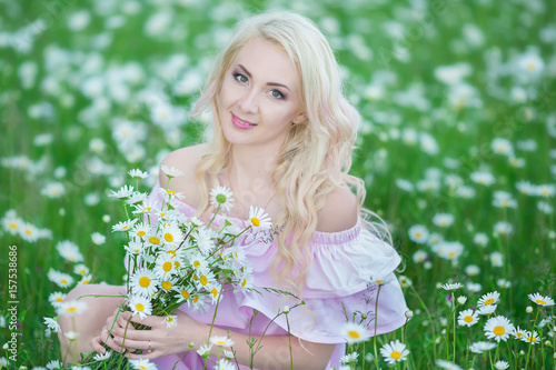 Attractive blonde in chamomile field. Young woman in wreath surrounded by chamomiles wearing stylish pink dress enjoying summer spring