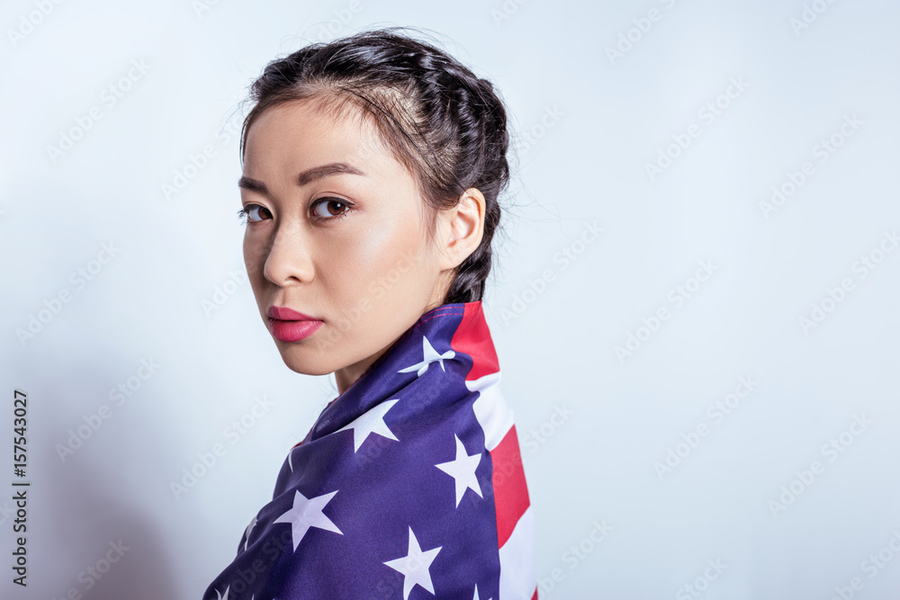 Portrait of stylish asian girl wrapped in american flag looking at camera isolated on grey Portrait of stylish asian girl wrapped in american flag looking at camera isolated on grey
