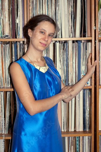 Young girl of 20 years in blue dress is standing near rack with old vinyl records and looking at camera.