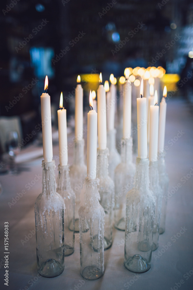 White candles in bottles of wine stand on white table