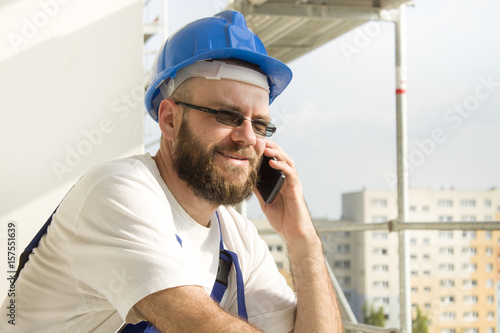 Obraz na płótnie Uśmiechnięty pracownik budowlany w stroju roboczym i w kasku na głowie rozmawia przez telefon. Praca na dużej wysokości. Rusztowania w tle. w salonie