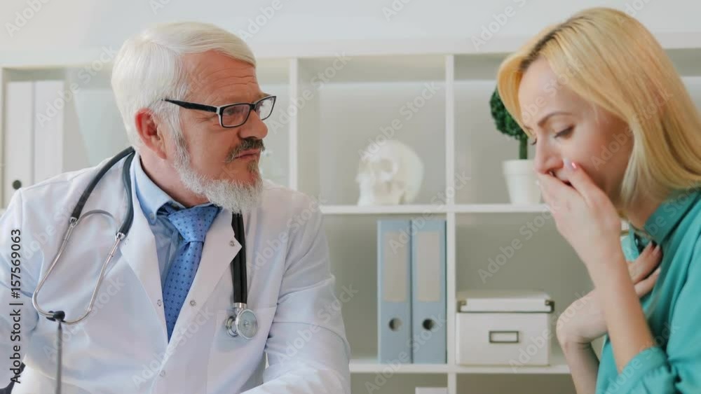 Coughing sick female patient complaining to caucasian midle aged doctor in glasses sitting in office about her throat. Coughing sick female patient complaining to caucasian midle aged doctor in glasses sitting in office about her throat.