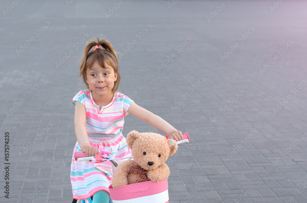 A beautiful little girl is riding a bicycle with a teddy bear in the basket. A beautiful little girl is riding a bicycle with a teddy bear in the basket.