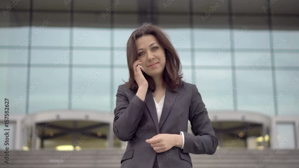 The girl assistant manager talking on the mobile phone with his colleague and looking at the camera. In the background the entrance to the business center. UHD 4K The girl assistant manager talking on the mobile phone with his colleague and looking at the camera. In the background the entrance to the business center. UHD 4K