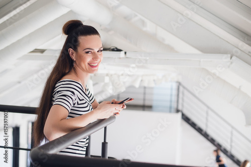 Happy woman smiling and standing indoors using a smartphone and looking at camera laughing.