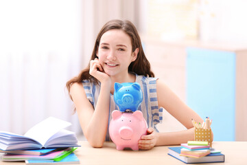 Smiling girl sitting at table with piggy banks and stationery. Saving for education concept