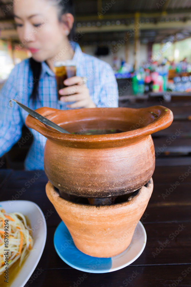 Baked clay pot of hot soup or Asian food on wooden table with background women hold glass of soda