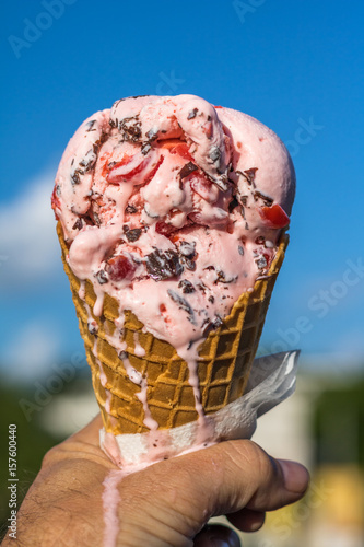 Vertical close up of melting cherry chocolate chip ice cream in a waffle cone being held in a hand with a bright blue sky with white clouds in the background
