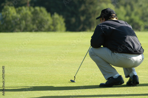 Man squats down on the golf course analyzing his shot. Man putting on the golf course. One man golfing on a sunny day in Utah wearing blue long sleeves shirt and khaki trousers. Serious golfer.