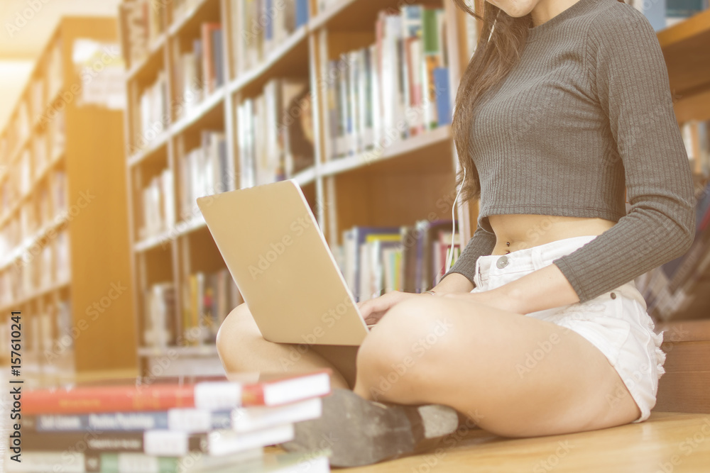 Obraz premium Asian sexy young girl student sit on the floor searching the data by laptop and wear earplug at bookshelf in library