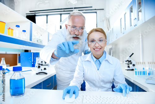 Smiling scientists in protective eyeglasses working together in lab and pointing at camera