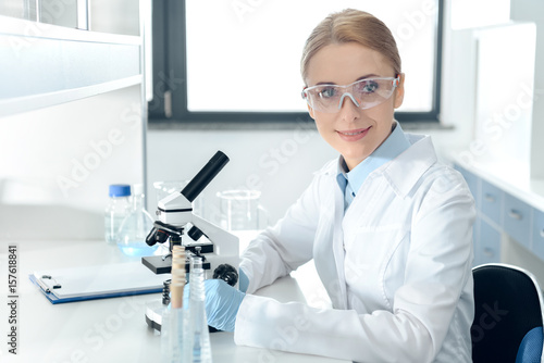 Smiling professional scientist in protective goggles working with microscope in chemical lab