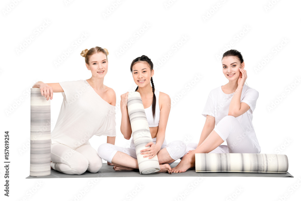 happy multiethnic girls resting after yoga training and sitting with yoga mats and looking at camera isolated on white