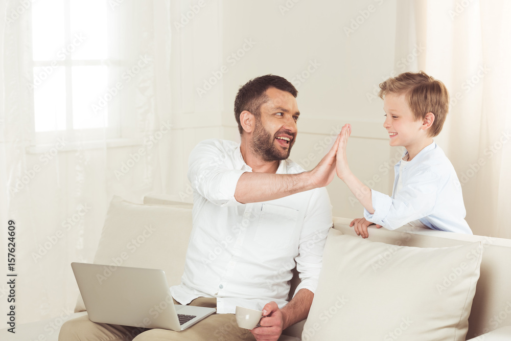 father sitting with laptop and giving high five to his little son at home father sitting with laptop and giving high five to his little son at home