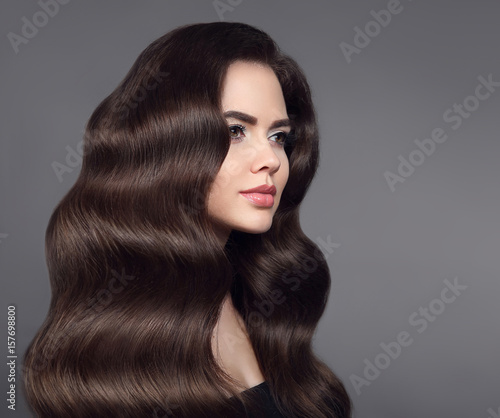 Healthy hair. Brunette girl portrait with long shiny wavy hair. Beautiful model with curly hairstyle and makeup isolated on studio dark background. Shampoo care product.