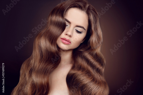 Beauty hair. Brunette girl with long shiny wavy hair. Beautiful model portrait with curly hairstyle and eyeliner makeup isolated on studio dark background.