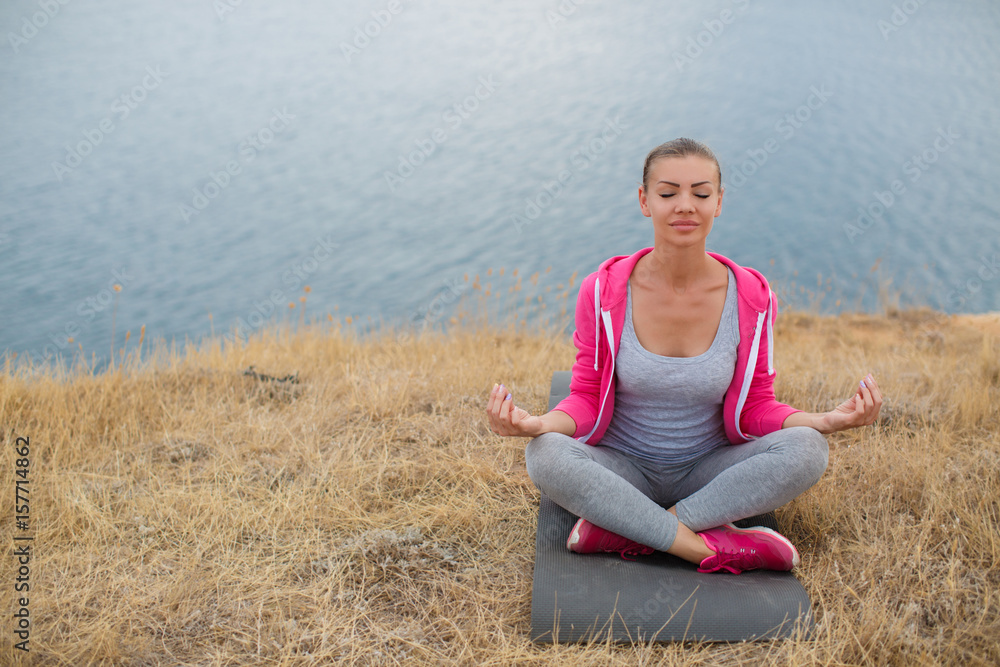 Athletic woman,brunette,hair in a braid,wearing a gray t-shirt and grey sweat pants,a pink sports jacket,performs relaxation exercises, sitting on a rocky beach near the sea in the Lotus position