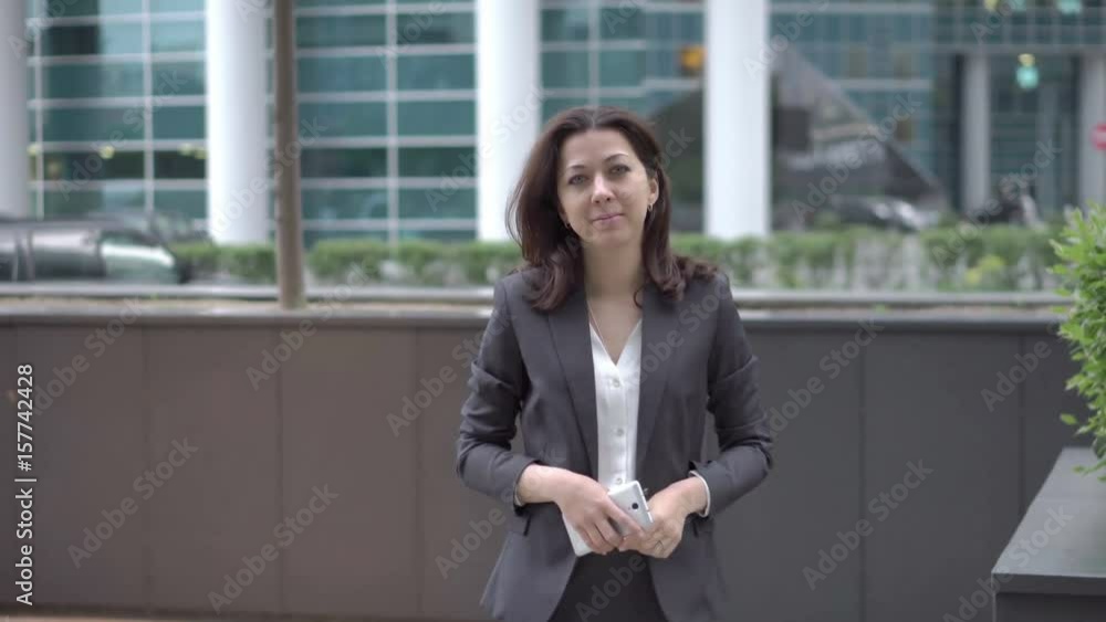 Attractive woman in business clothes with a mobile phone in hand approaching to the camera, stops and smiles. She is on the background buildings business centers, UHD 4K Attractive woman in business clothes with a mobile phone in hand approaching to the camera, stops and smiles. She is on the background buildings business centers, UHD 4K
