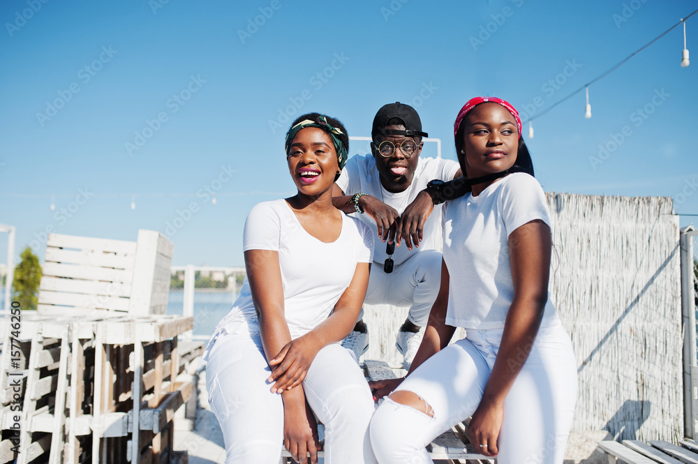 Three stylish african american friends, wear on white clothes at pier on beach. Street fashion of young black people. Black man with two african girls.