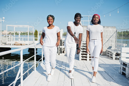Three stylish african american friends, wear on white clothes at pier on beach. Street fashion of young black people. Black man with two african girls. © AS Photo Family