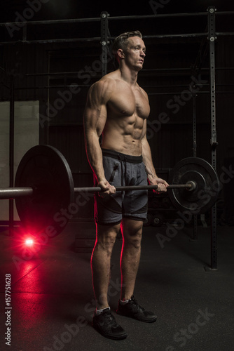 Muscular White Caucasian man doing a barbell bicep curl in a dark grungy gym wearing shorts and showing muscular body and six pack abs 